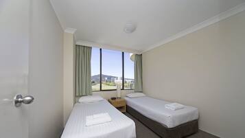 Habitación de hotel con dos camas individuales, una mesita de noche, una lámpara y ventana con vistas a la ciudad y montañas.