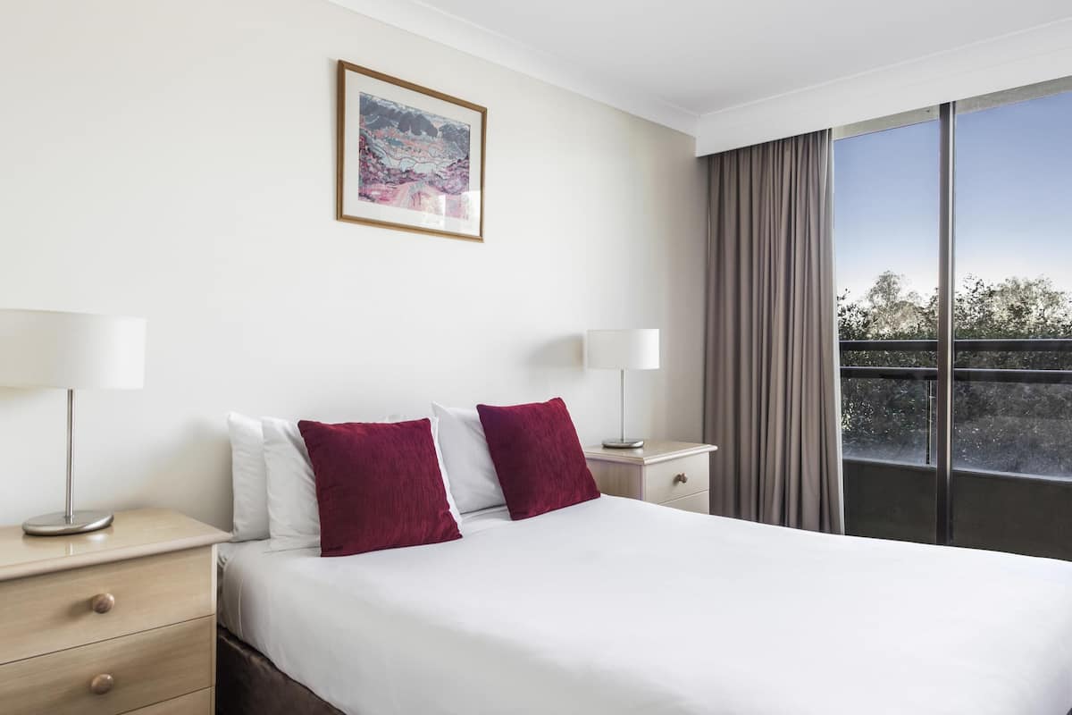 A hotel room with a large bed, two bedside tables, a nightstand, a framed picture on the wall, and a view of the outdoors through a window.