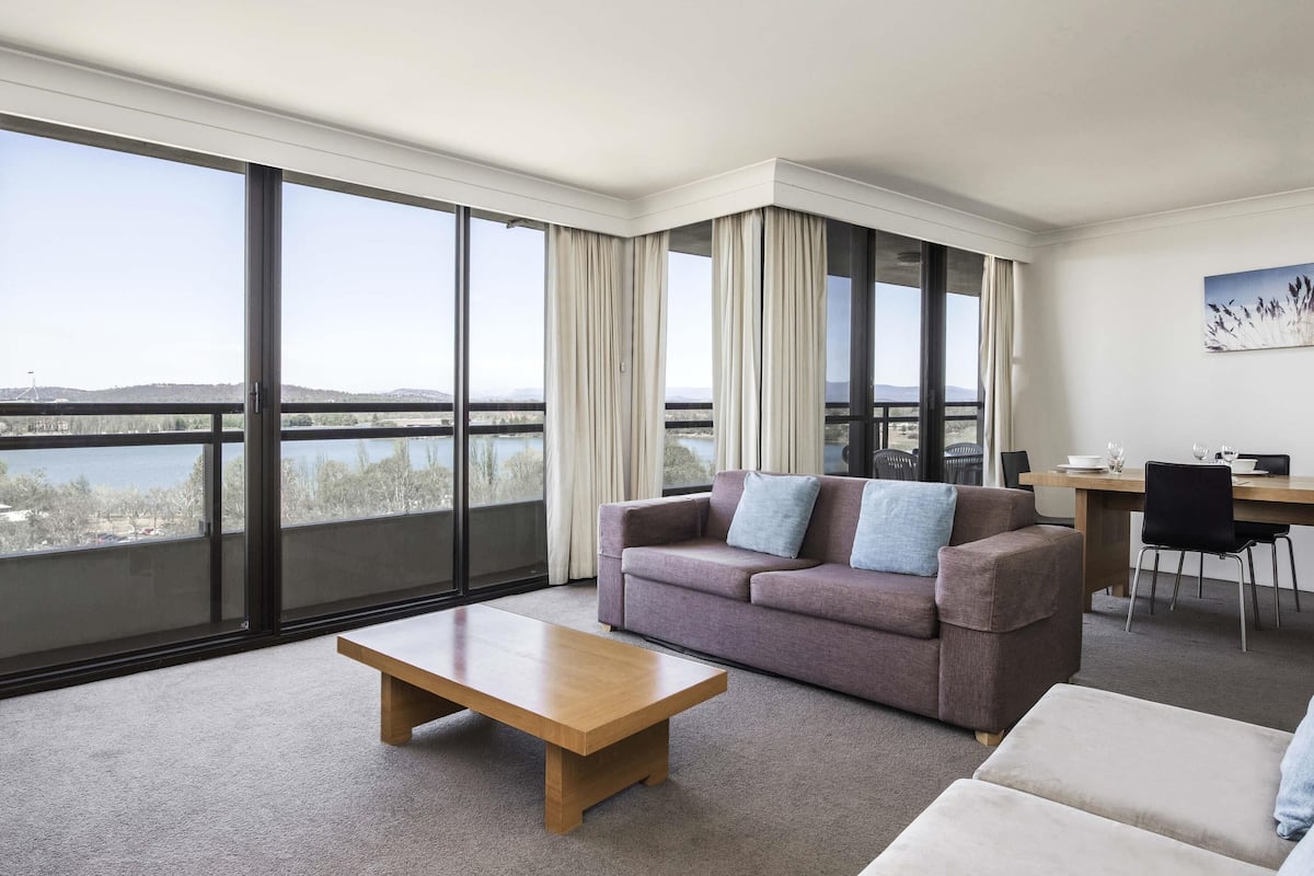 A modern hotel room with a large balcony, a sofa, a coffee table, and a dining area.