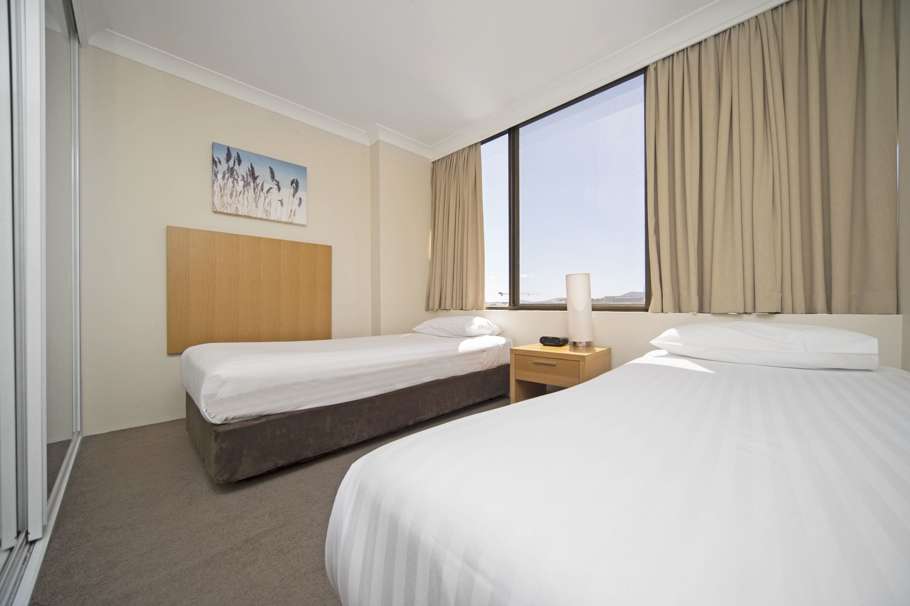 Habitación de hotel con dos camas individuales, cabecera de madera, mesita de noche con lámpara y un amplio ventanal con cortinas.