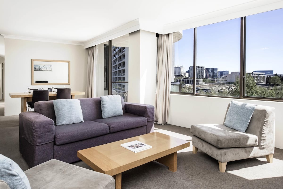 A modern hotel room with a large window, a sofa, a chair, a coffee table, and a dining area.