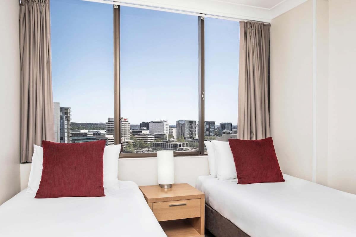 A hotel room with two beds, a nightstand, and a view of a cityscape.