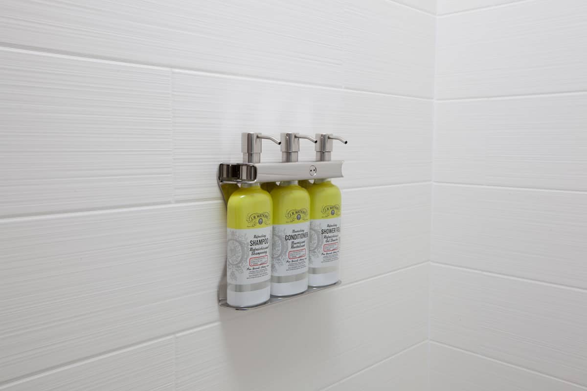 A bathroom with white tiled walls and a shelf holding three bottles of shampoo and conditioner.