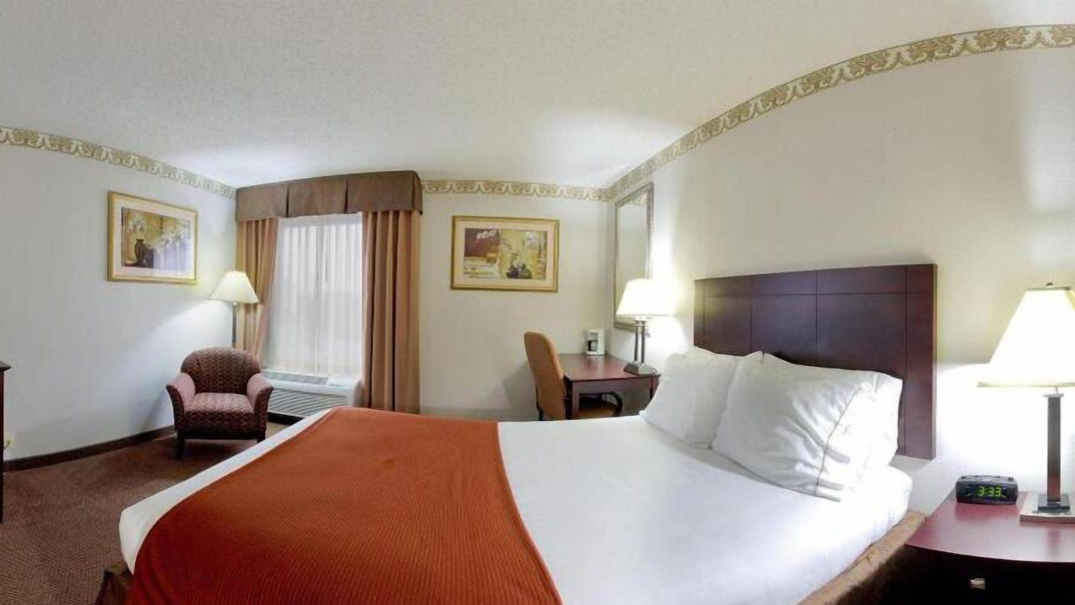 A hotel room with a large bed, a desk, a chair, a window with curtains, and two wall-mounted lamps.