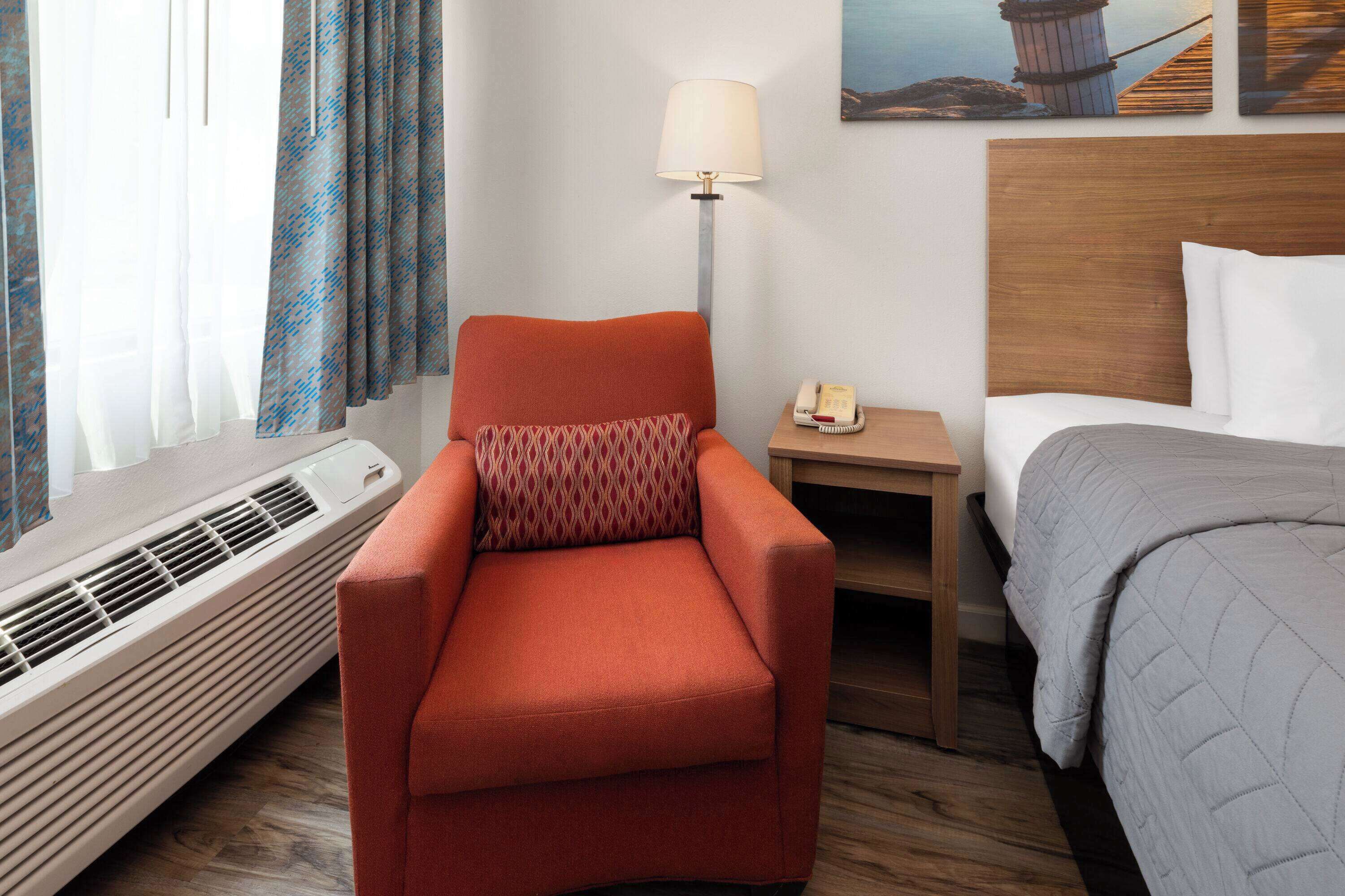 Une chambre d’hôtel avec un fauteuil rouge, une table de chevet avec une lampe, un lit recouvert d’une couverture grise et un tableau accroché au mur.
