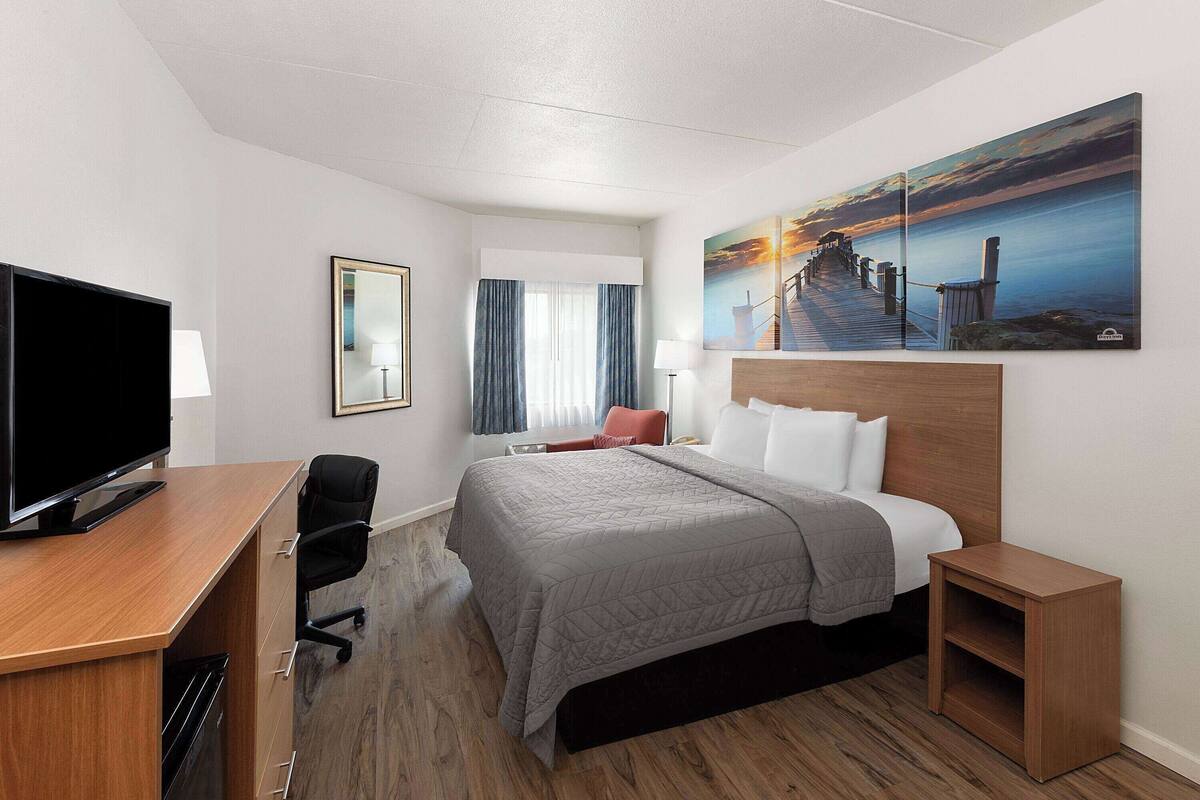 A hotel room with a bed, a desk with a TV, a chair, a nightstand, and a painting of a pier.