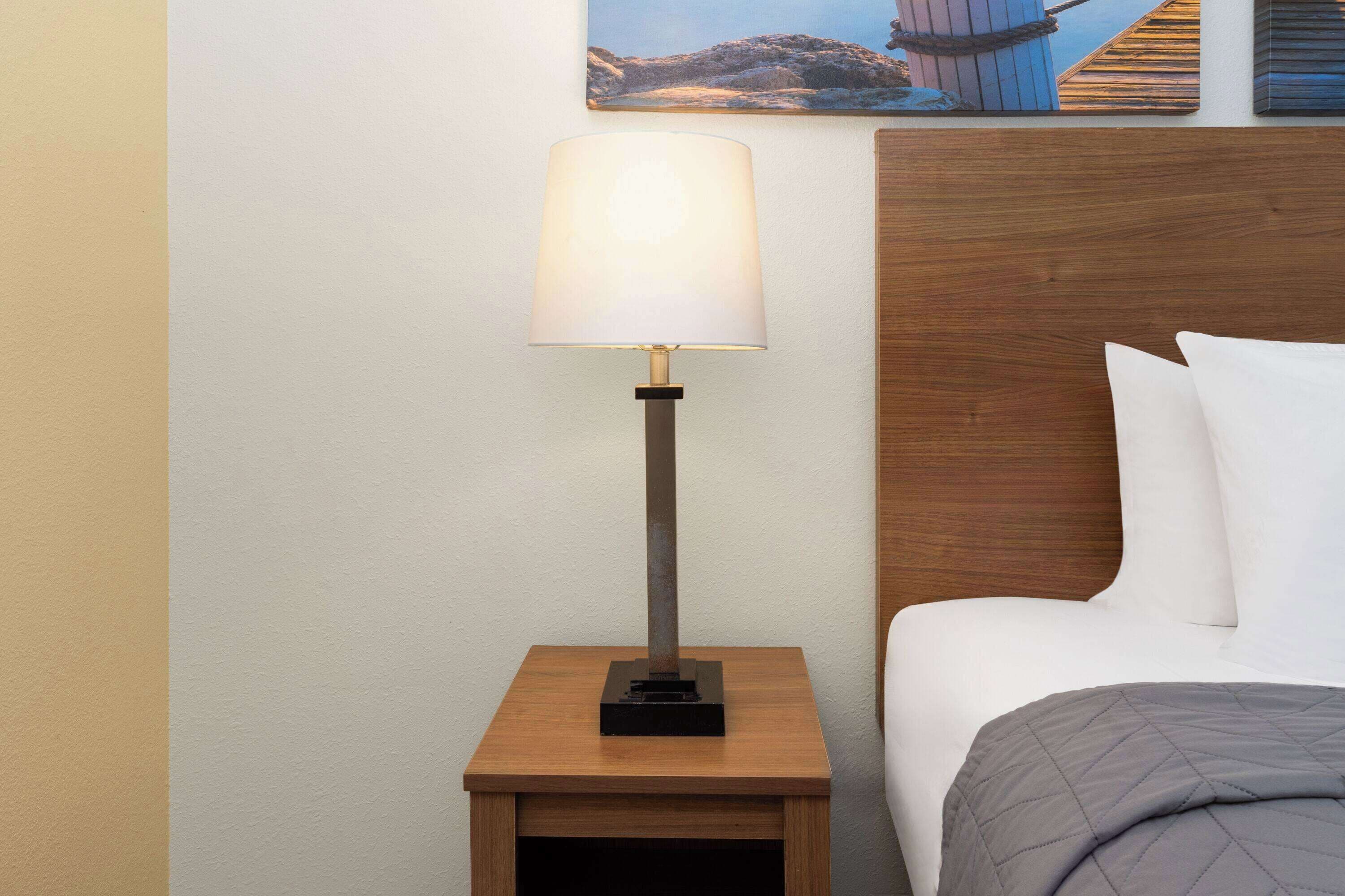 Een bed met een houten hoofdbord, een nachtkastje met een lamp en een schilderij aan de muur.