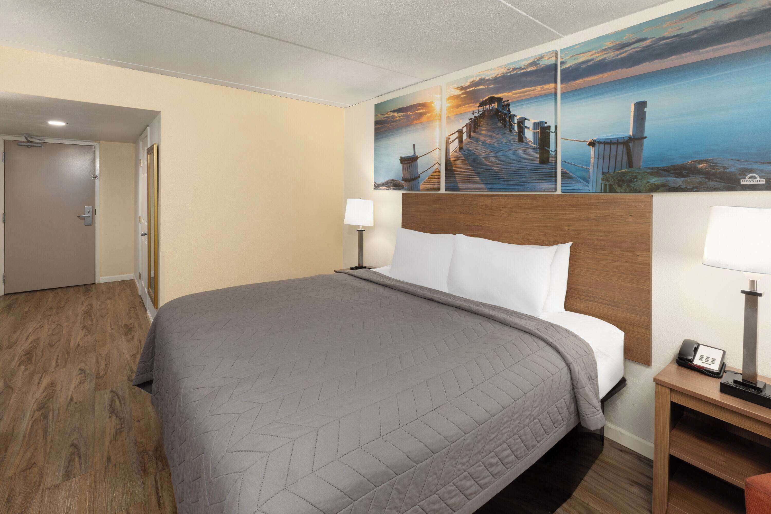 A hotel room with a large bed, a wooden headboard, a nightstand with a lamp, and a painting of a pier on the wall.