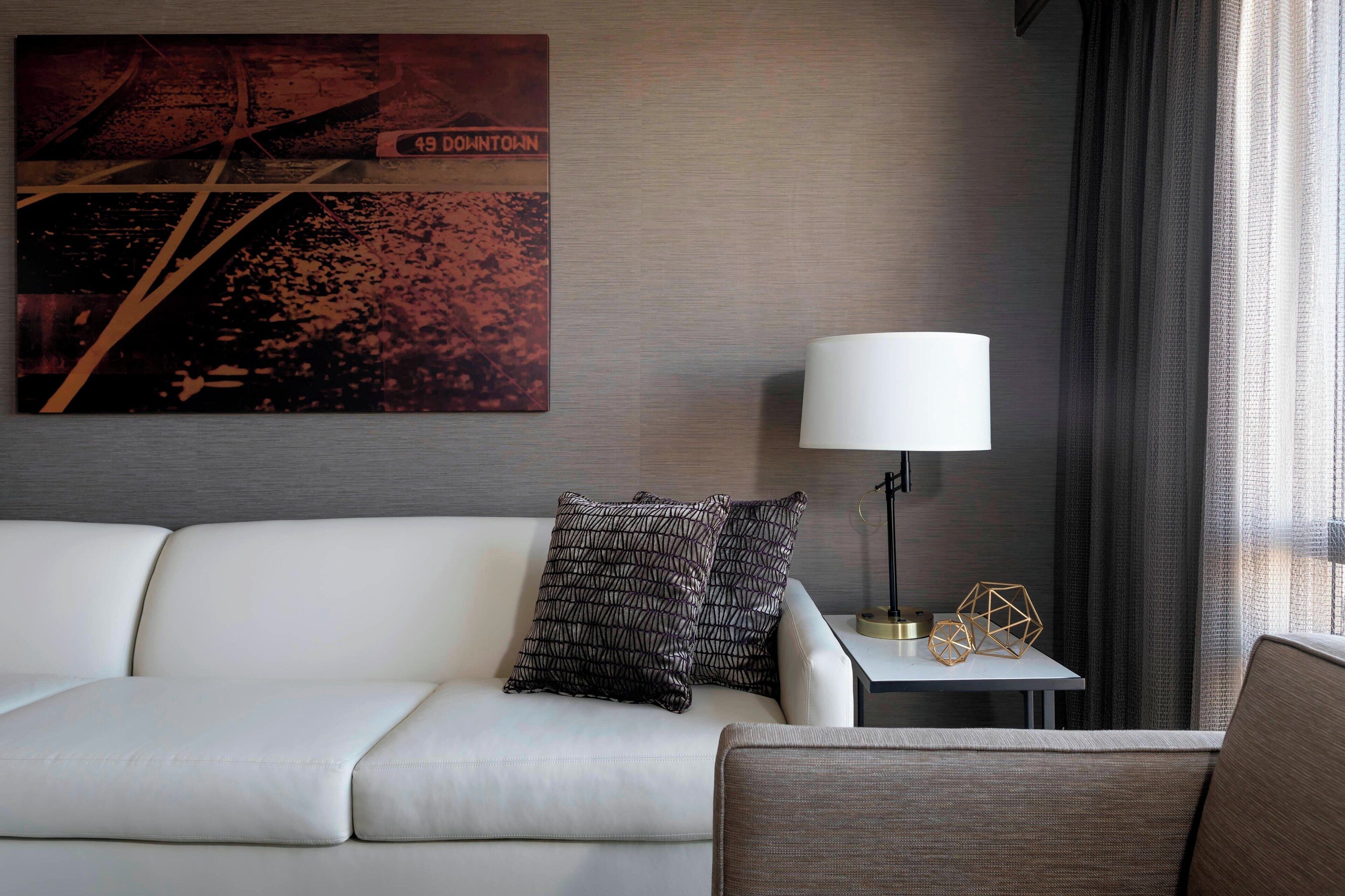 A modern hotel room with a white sofa, a side table with a lamp, decorative geometric objects, and a cityscape artwork on the wall.