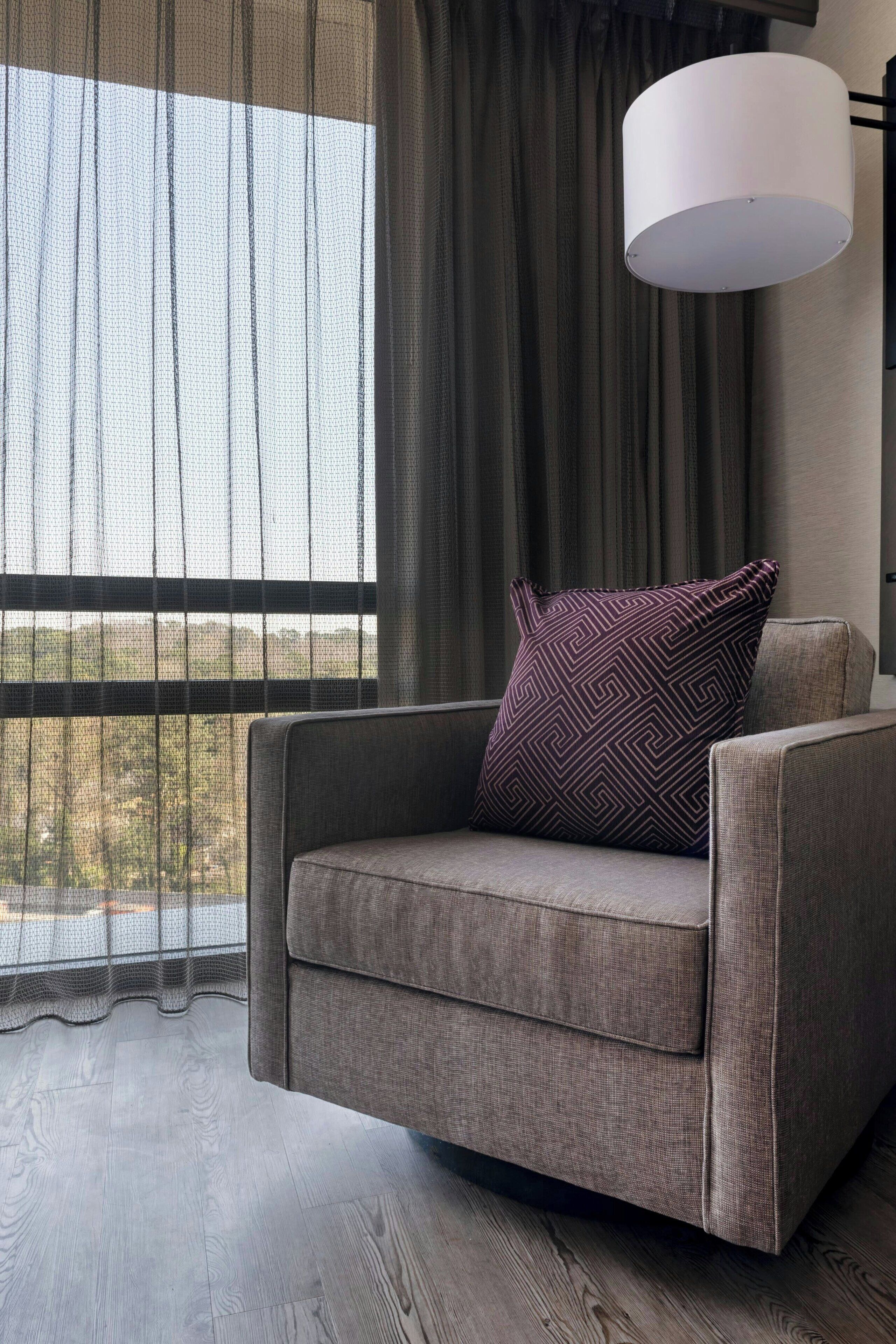 A modern living room with a grey sofa, a patterned throw pillow, a large white floor lamp, and a view through sheer curtains.