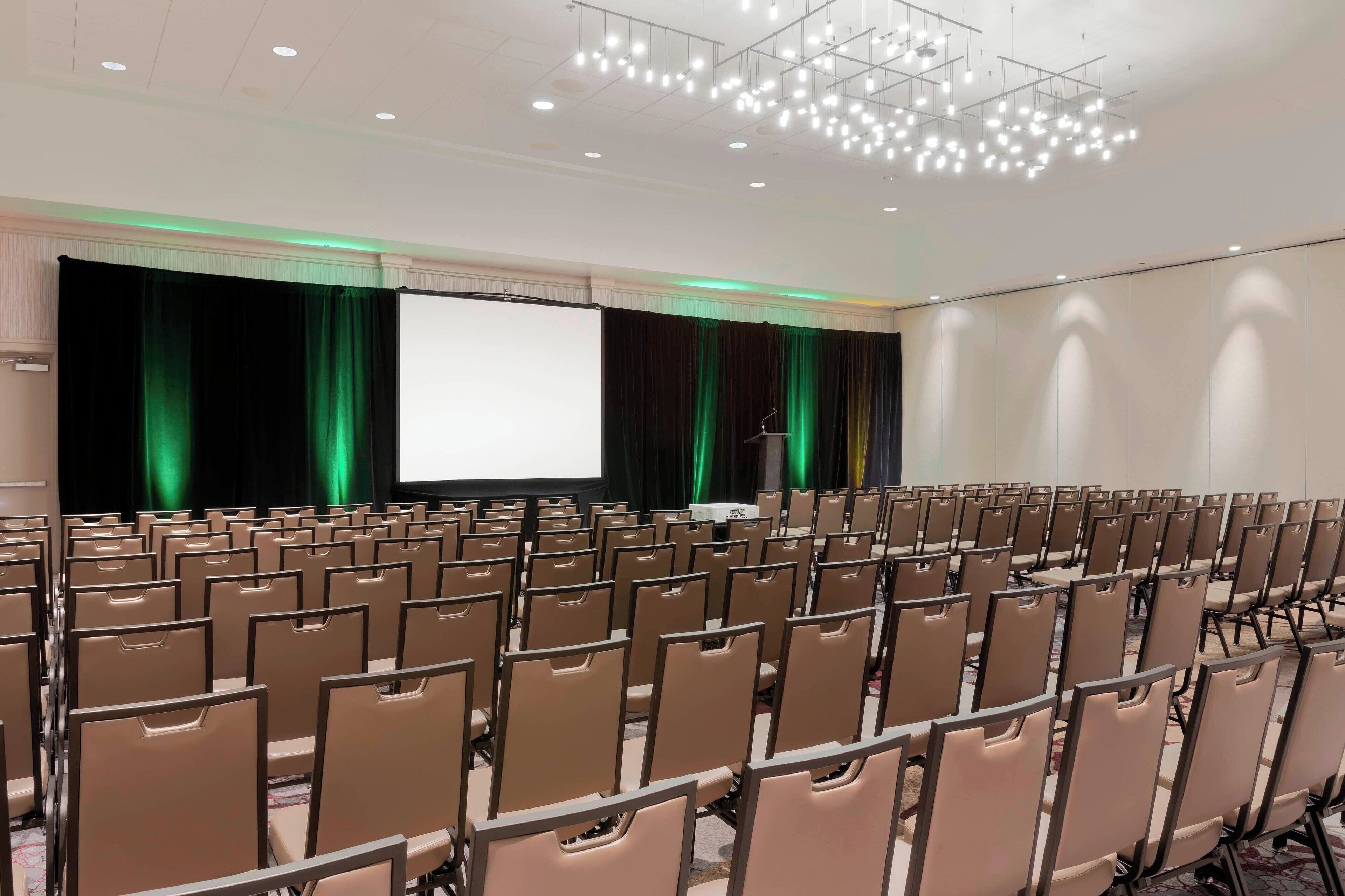 A conference room with rows of chairs, a stage with a projector screen, and a podium.