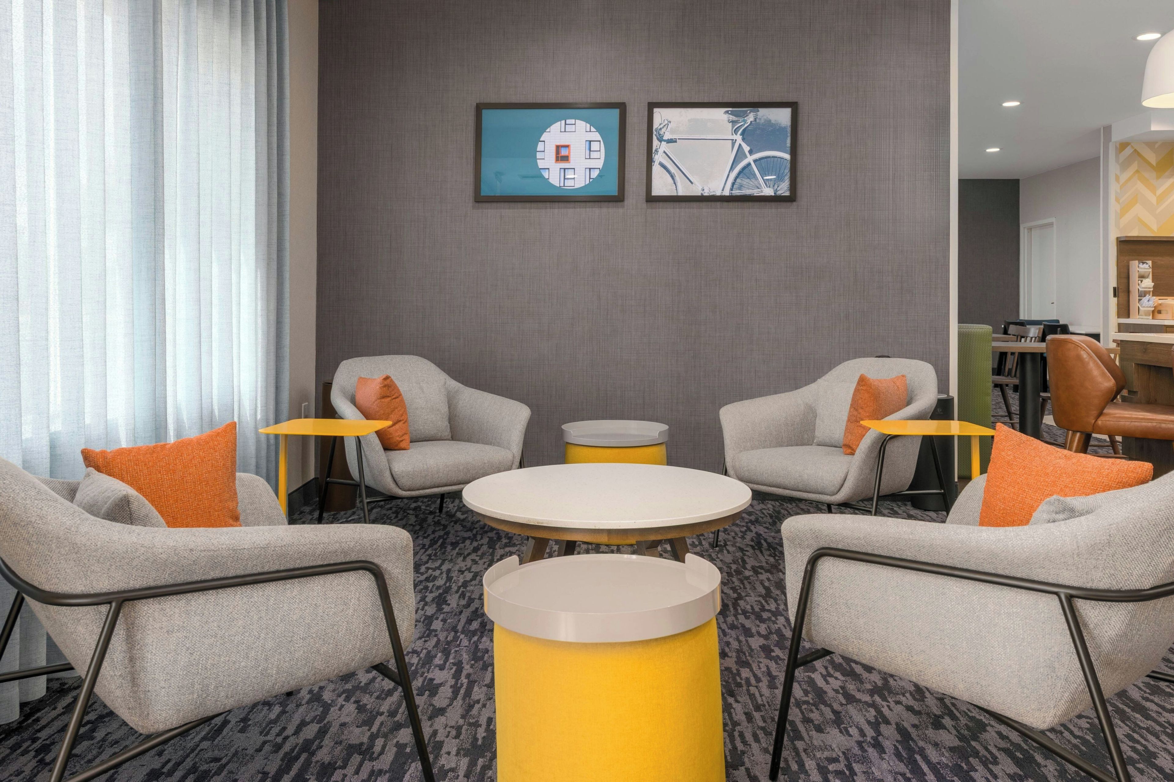 Un hall d’hôtel avec des canapés gris, des tables jaunes et deux œuvres d’art encadrées accrochées au mur.