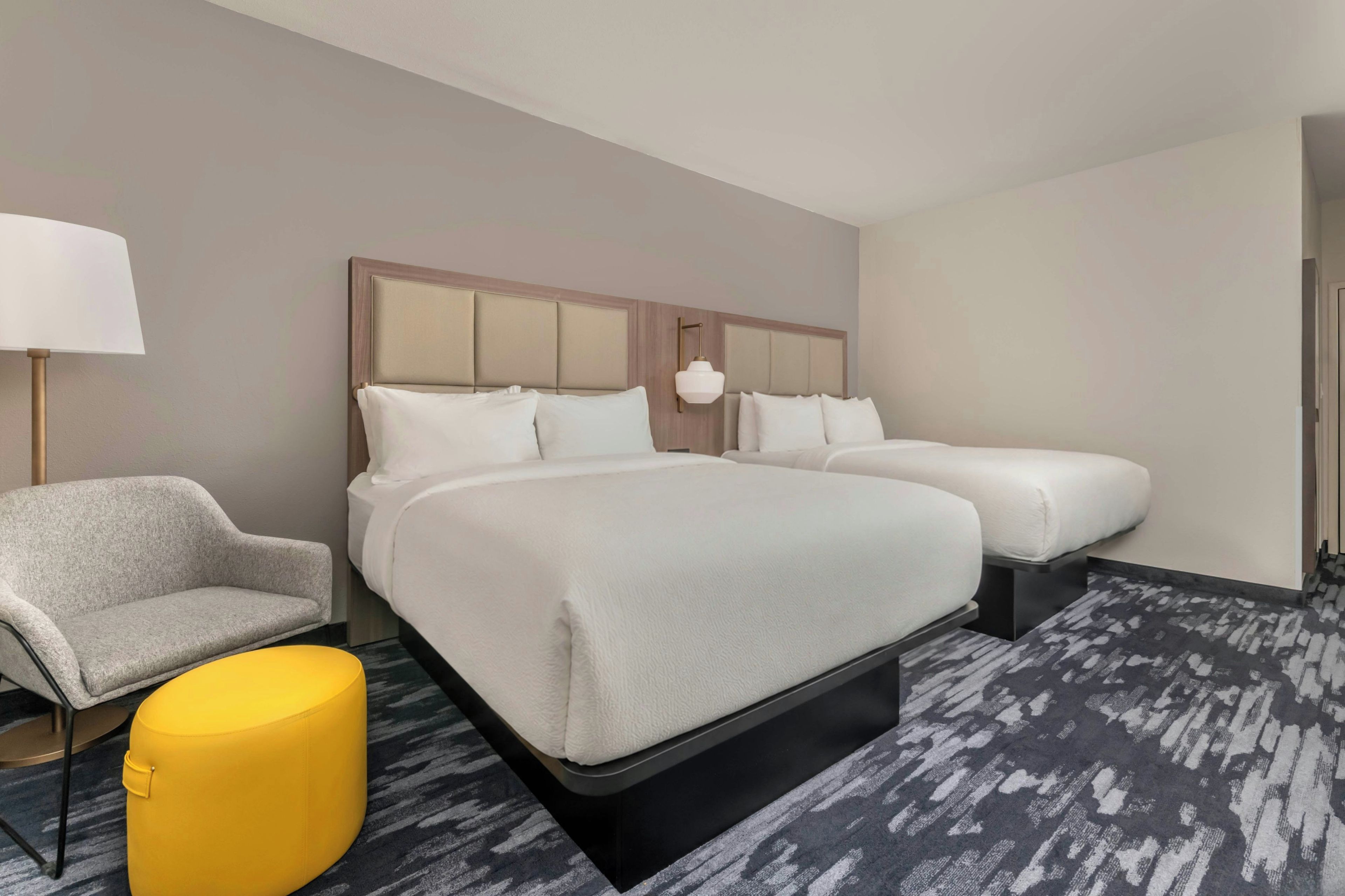 Une chambre d’hôtel avec deux lits, un canapé, un pouf jaune et une lampe.