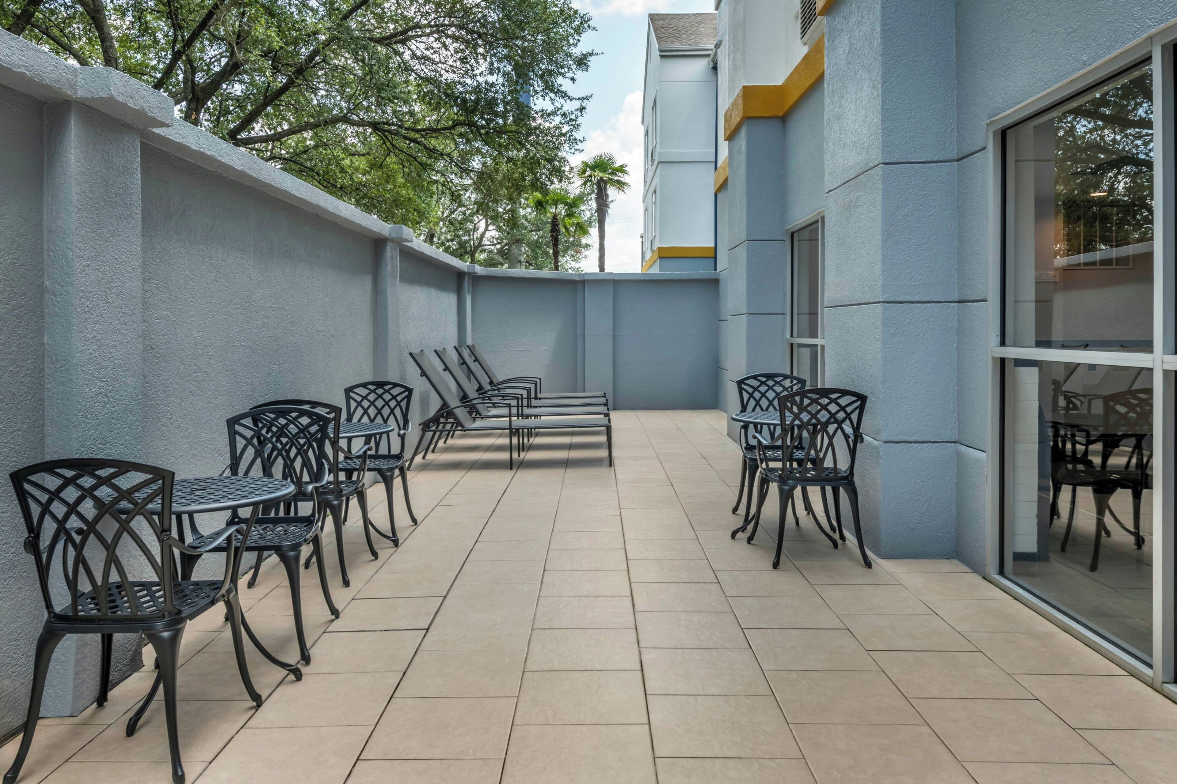 Un patio al aire libre con varias mesas y sillas, piso de baldosas y un edificio con grandes ventanales.
