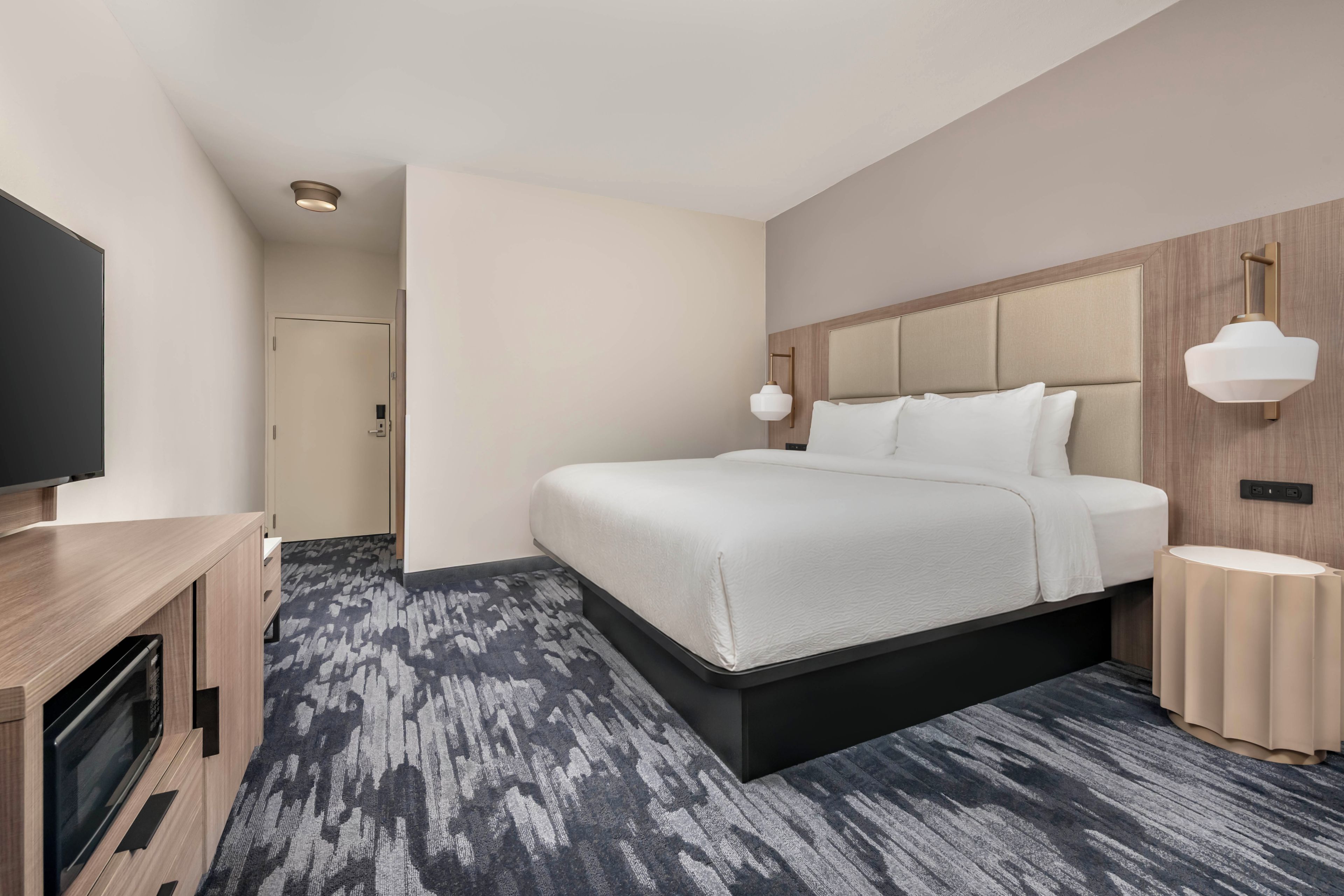 A modern hotel room with a large bed, a TV, a desk, and a carpeted floor.