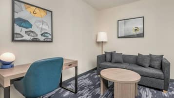 A modern hotel room with a desk, a blue office chair, a gray sofa, a round wooden coffee table, and two wall-mounted lamps.