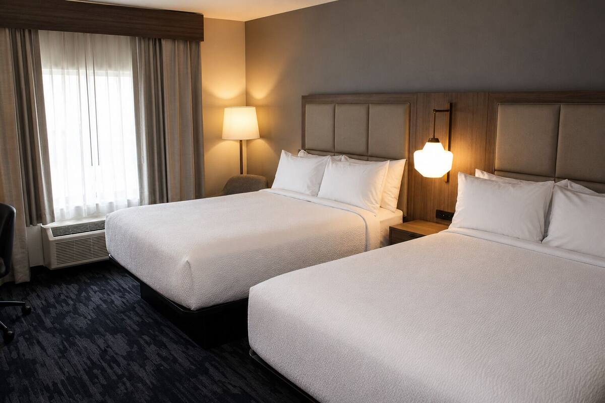 A hotel room with two beds, a desk, a chair, a window with curtains, and a wall-mounted lamp.
