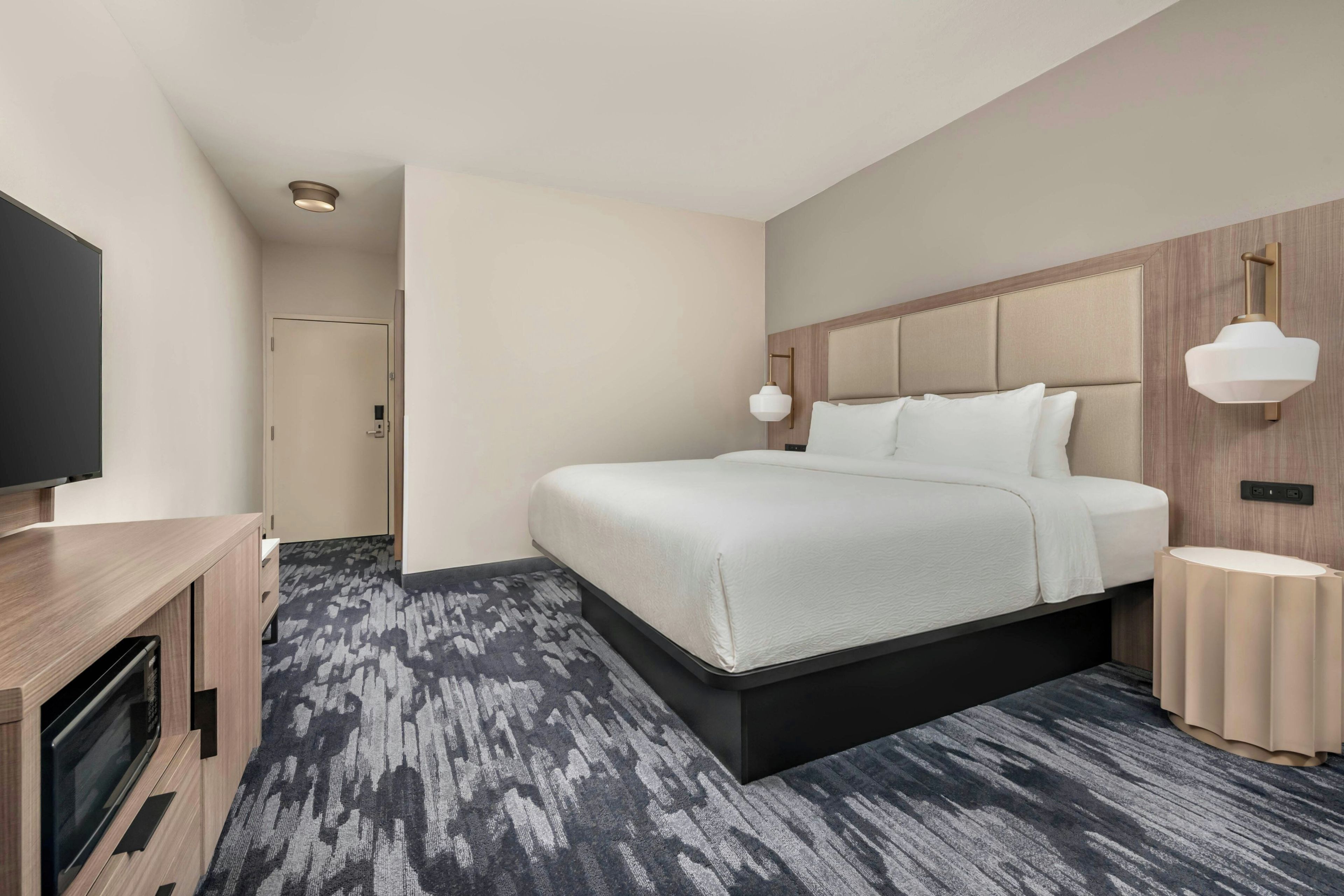 A modern hotel room with a large bed, a flat-screen TV, a wooden desk, and a patterned carpet.