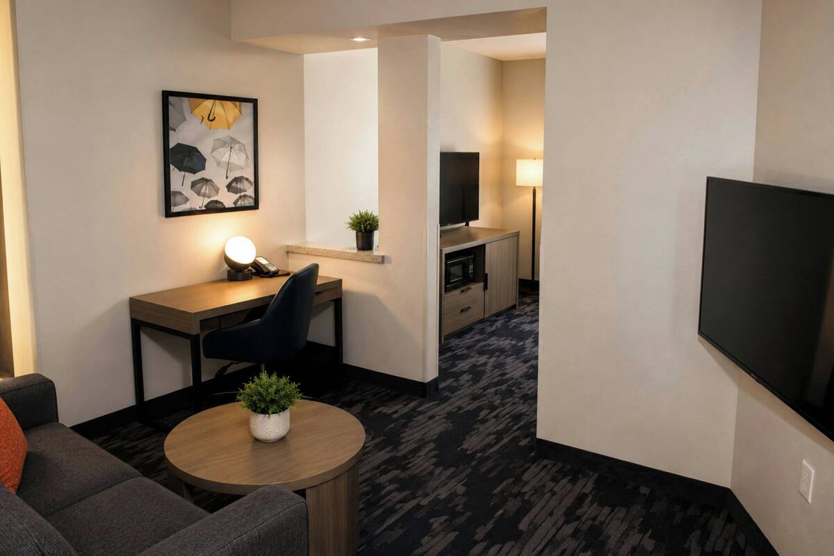 A hotel room with a sofa, a small round table with a plant, a desk with a chair, a TV, and a kitchen area with a microwave.