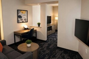 Une chambre d’hôtel comprenant un canapé, une petite table ronde avec une plante, un bureau avec une chaise, une télévision et un coin cuisine équipé d’un four à micro-ondes.