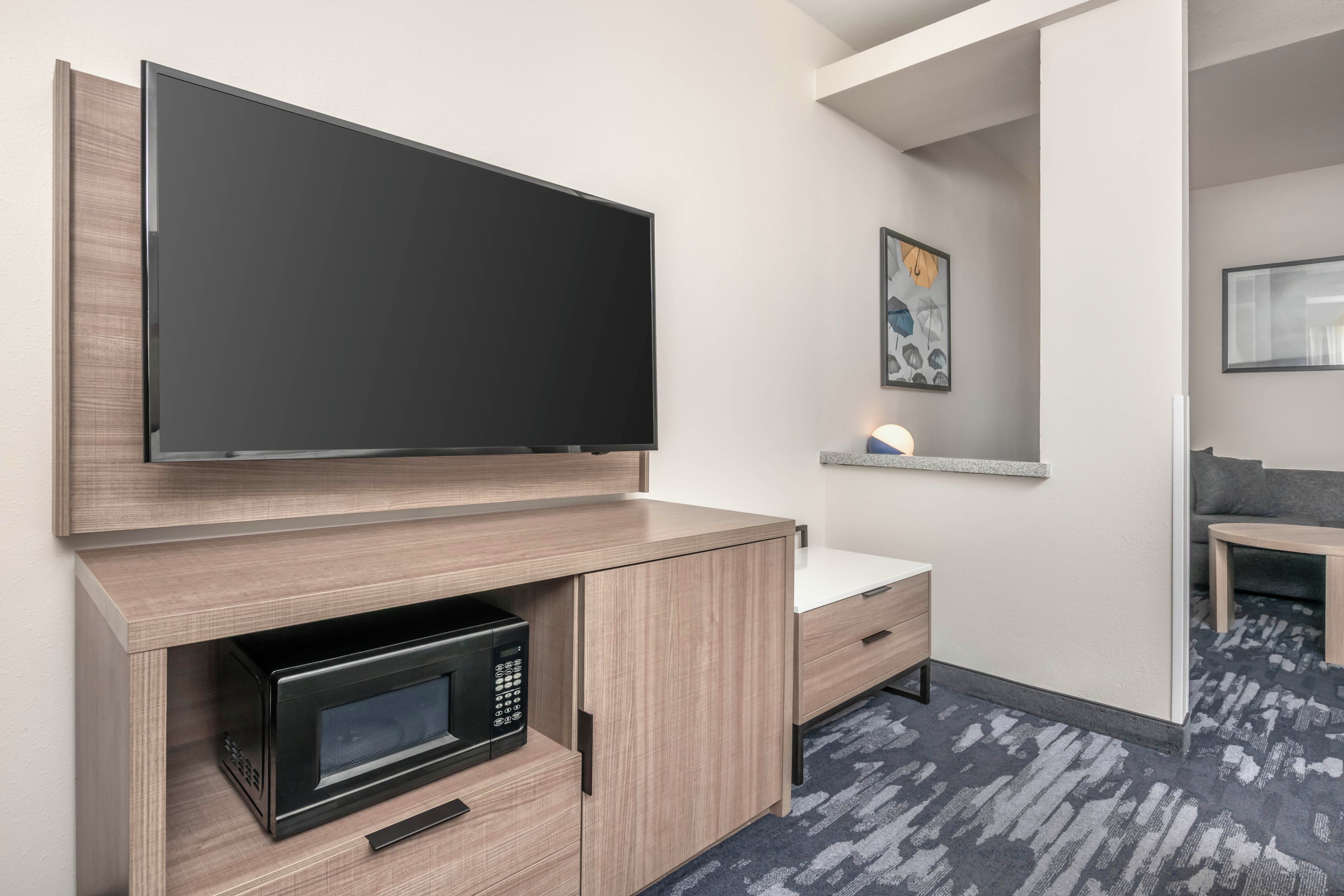A hotel room with a flat-screen TV mounted above a wooden cabinet, a microwave, and a small bedside table with a lamp.