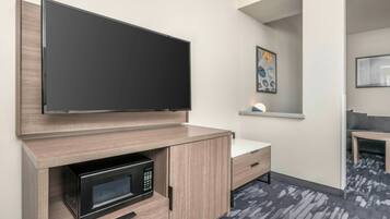 A hotel room with a flat-screen TV mounted on the wall, a built-in microwave, a small bedside table with a lamp, and a gray patterned carpet.