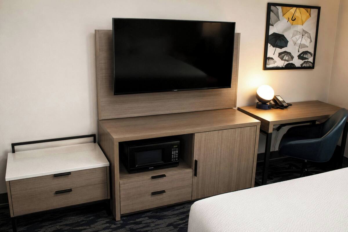 A hotel room with a flat-screen TV, a microwave, a desk with a chair, and a bedside table.