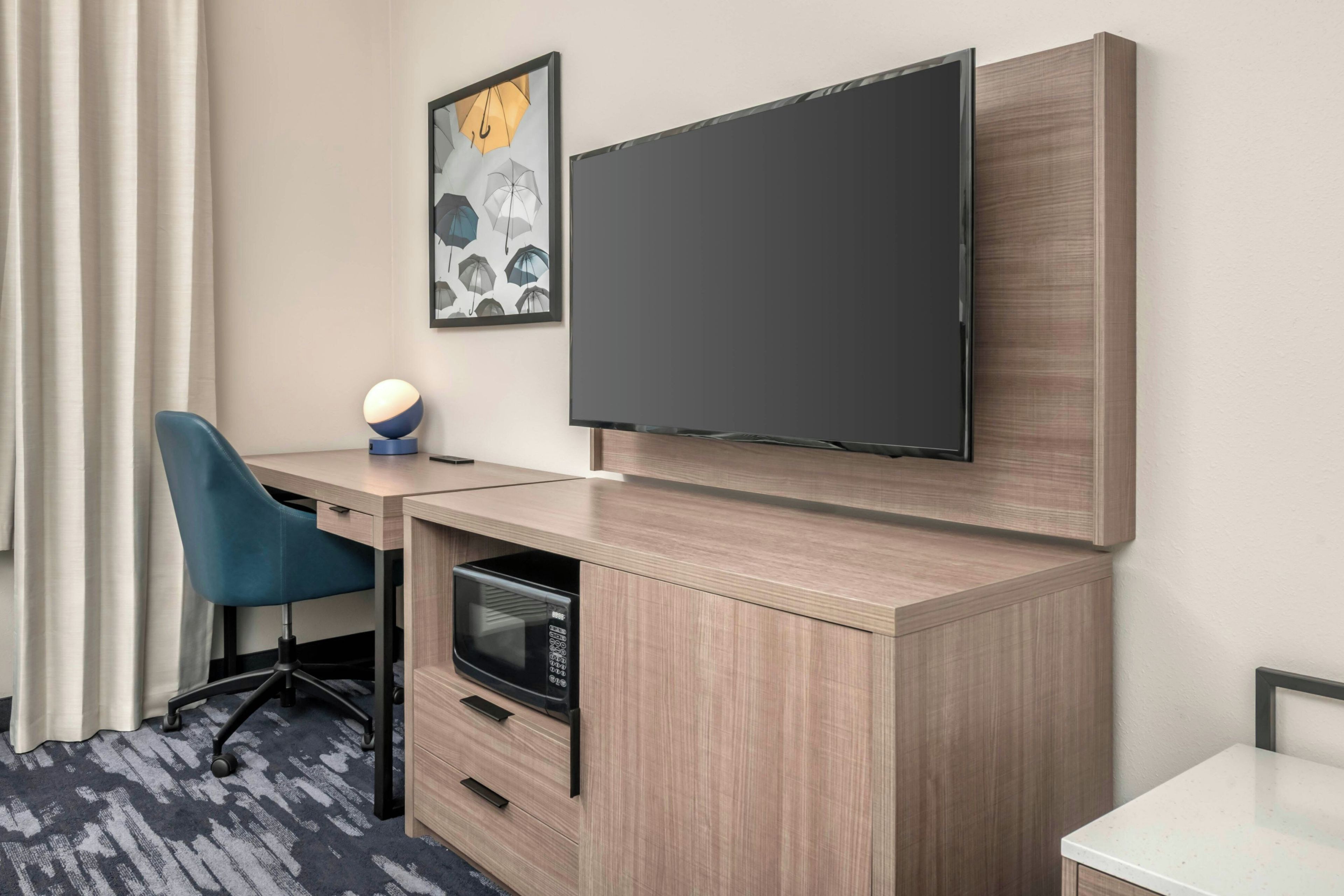 A hotel room with a flat-screen TV mounted on the wall, a desk with a chair, a microwave, and a small table.