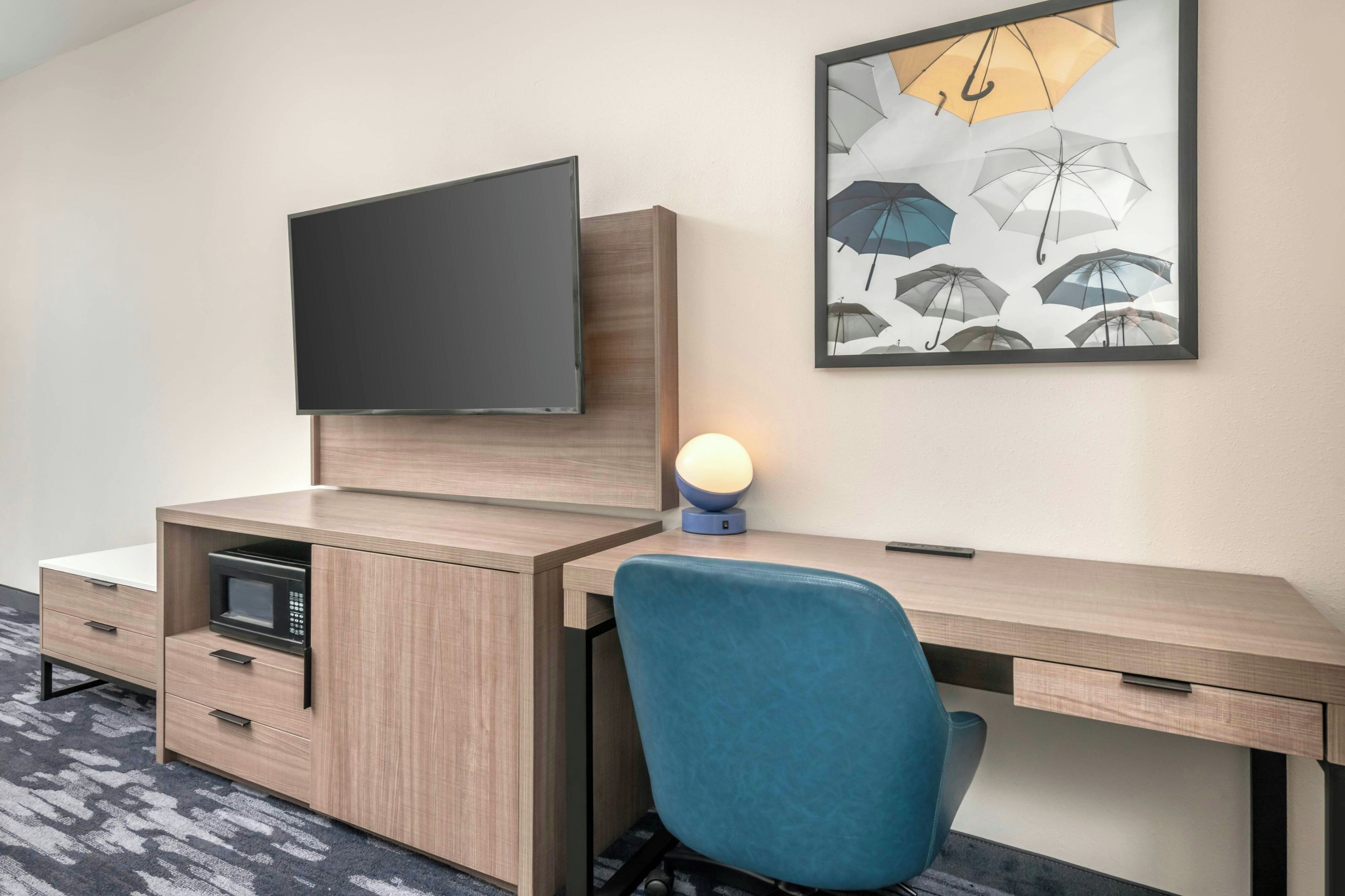 A hotel room with a flat-screen TV mounted on a wooden cabinet, a desk with a chair, and a wall-mounted lamp.