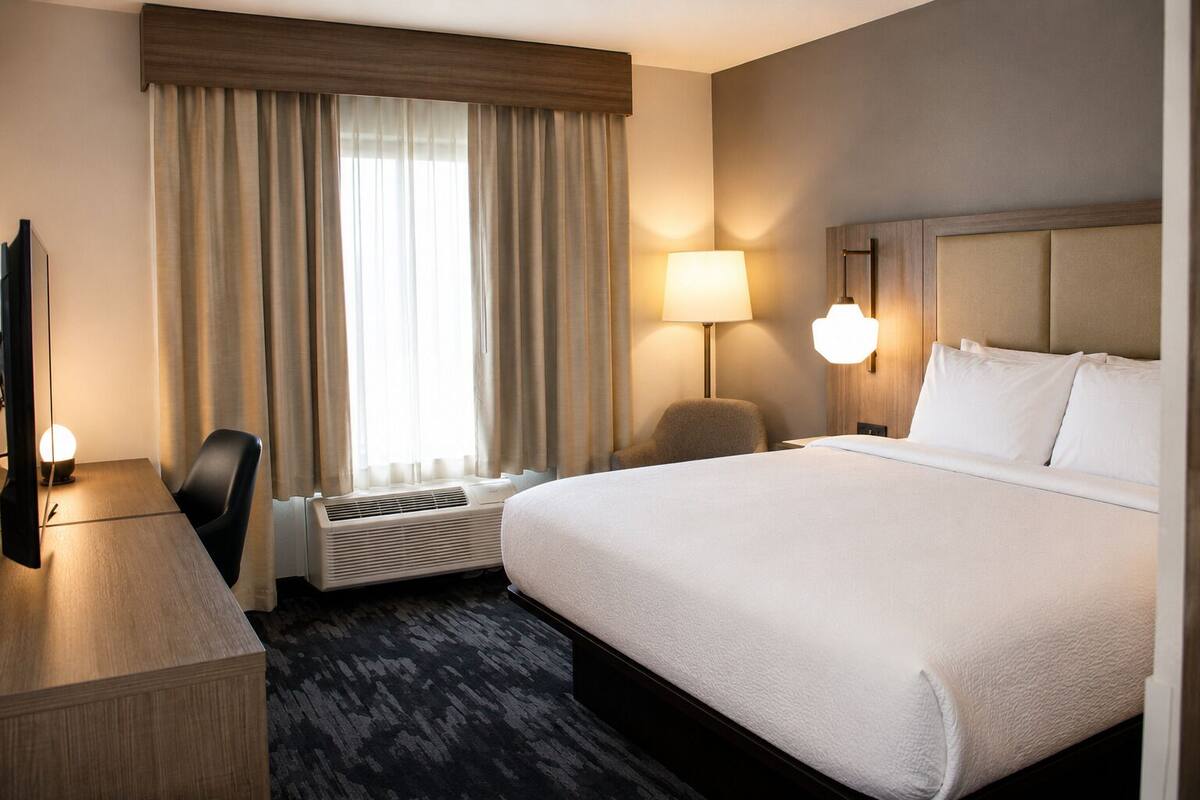 A hotel room with a bed, a desk, a chair, a lamp, and a window with curtains.