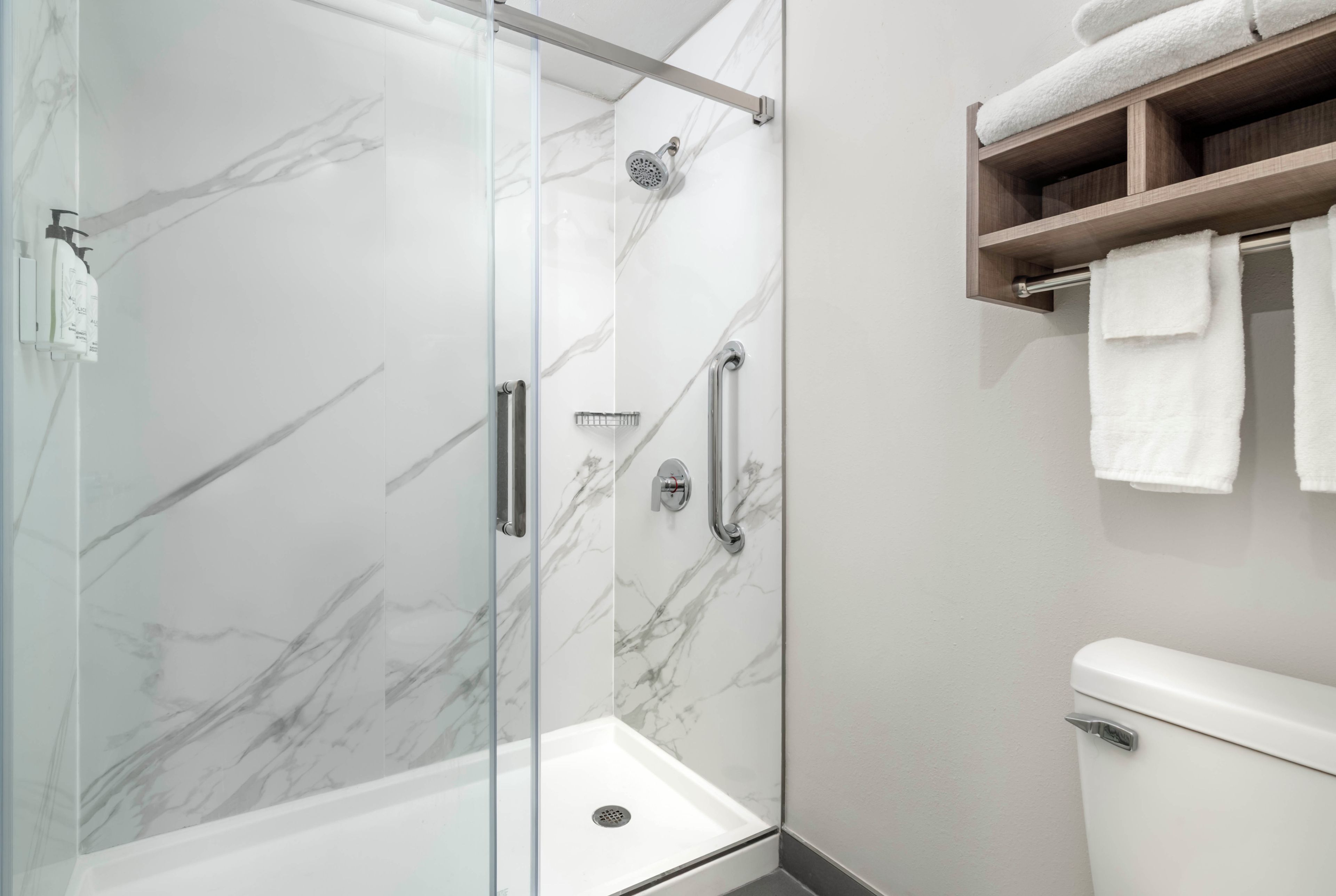 A modern bathroom with a glass-enclosed shower, marble walls, a toilet, and a shelf with towels.