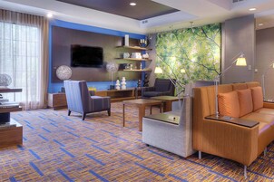 A modern hotel room with a blue and yellow patterned carpet, a comfortable sofa, a small table, a chair, a TV mounted on the wall, and a bookshelf with decorative items.