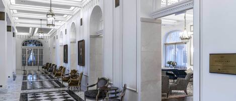 A hotel lobby with a checkered marble floor, elegant chairs, and chandeliers.