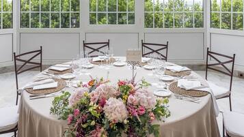 A round table set for a formal event with a floral centerpiece, white tablecloth, and dark wooden chairs.