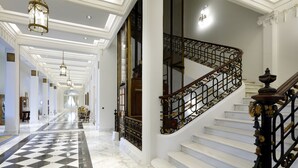 Un amplio pasillo con suelo de mármol a cuadros, una escalera con barandilla de metal negro ornamentada y un candelabro.