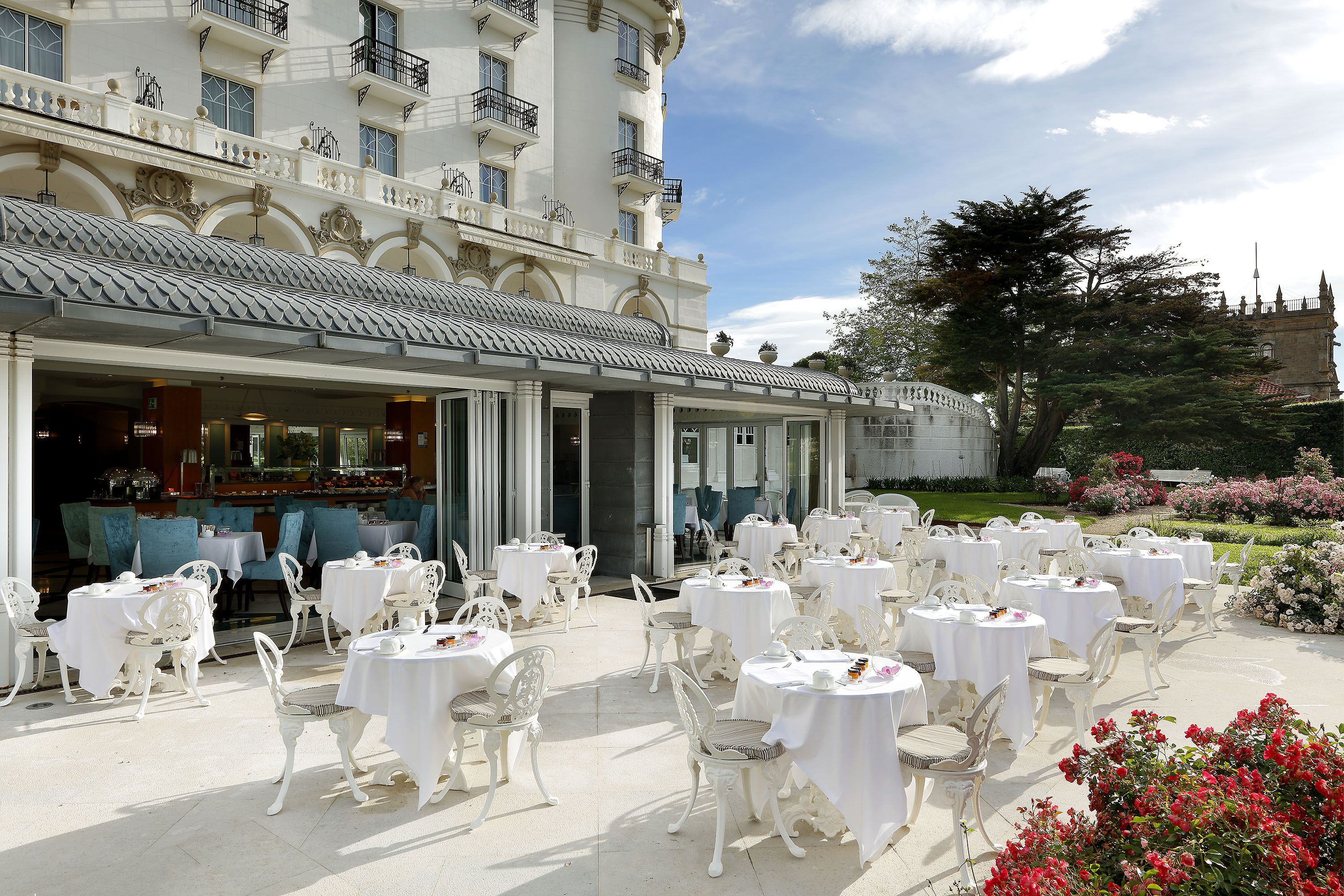 Un espace extérieur aménagé pour les repas, avec des tables et des chaises blanches, un grand arbre et, au fond, un bâtiment doté de balcons.