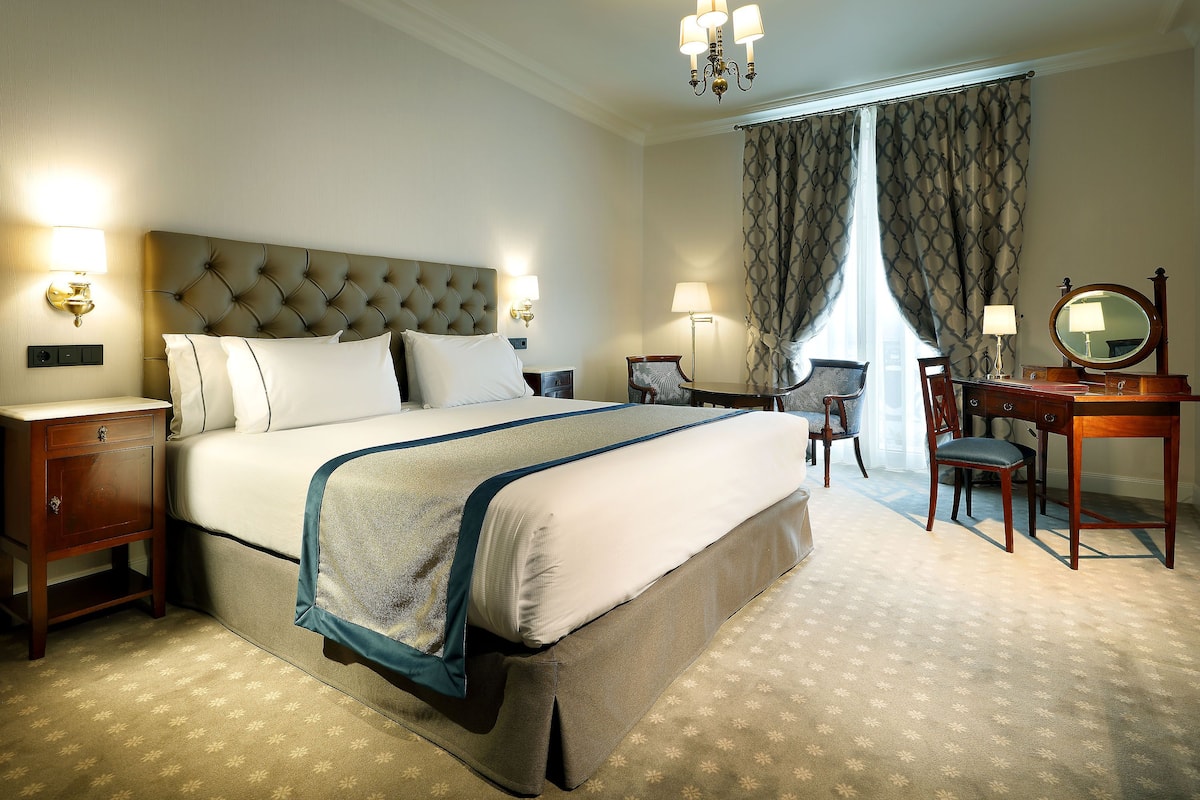 A neatly arranged hotel room with a large bed, bedside tables, a desk with a chair, and a mirror.