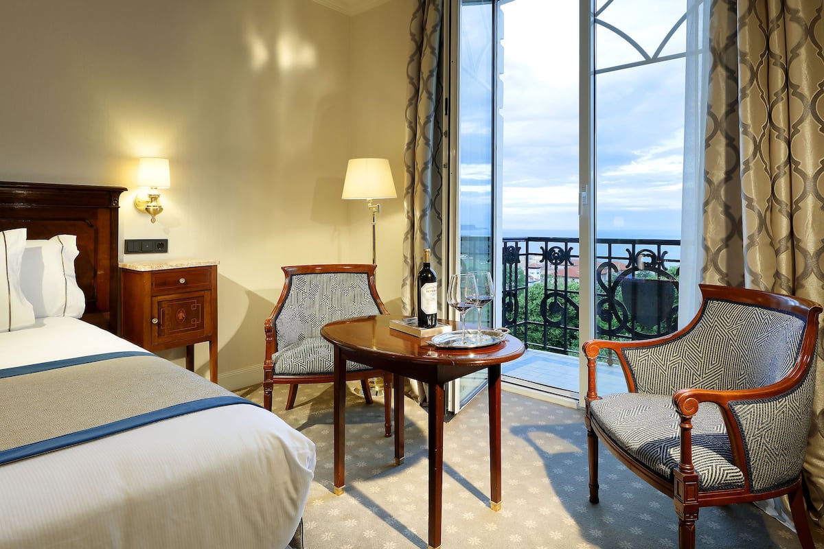 A hotel room with a bed, two chairs, a small table, and a balcony with a view.