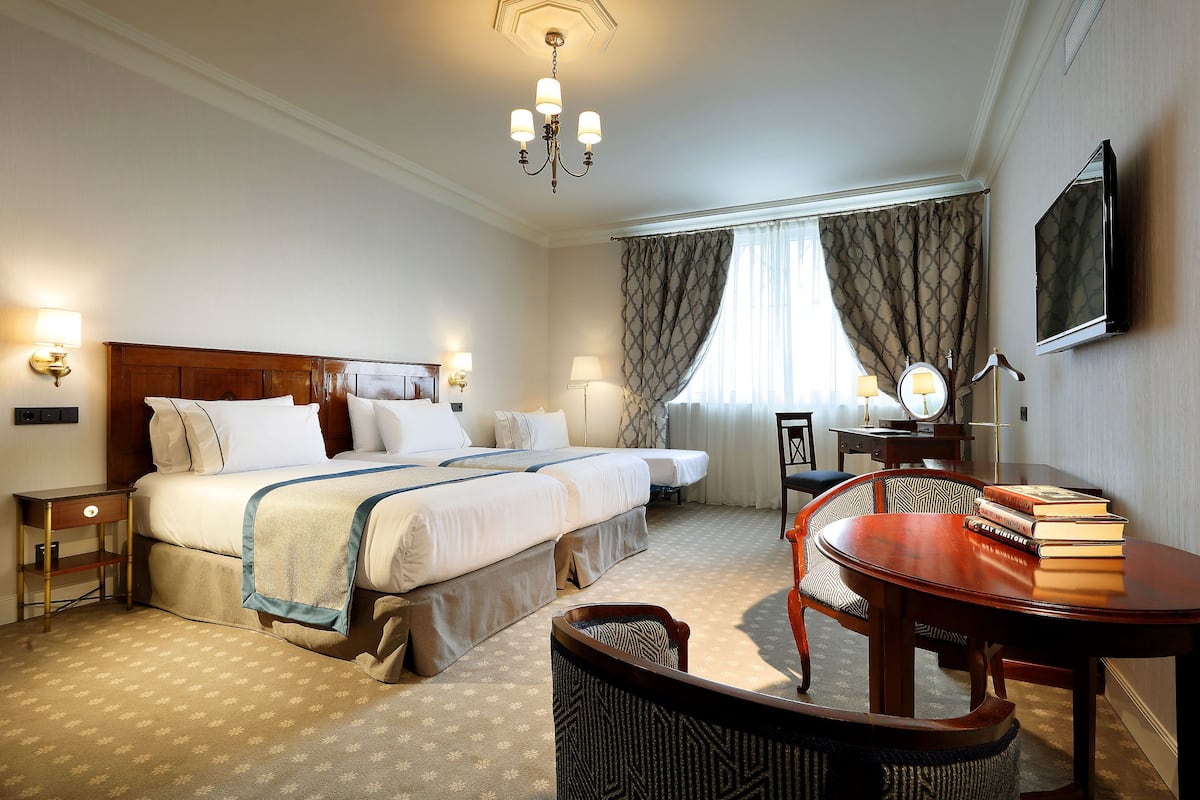 A hotel room with a double bed, bedside tables, a desk with a chair, a TV, and a chandelier.