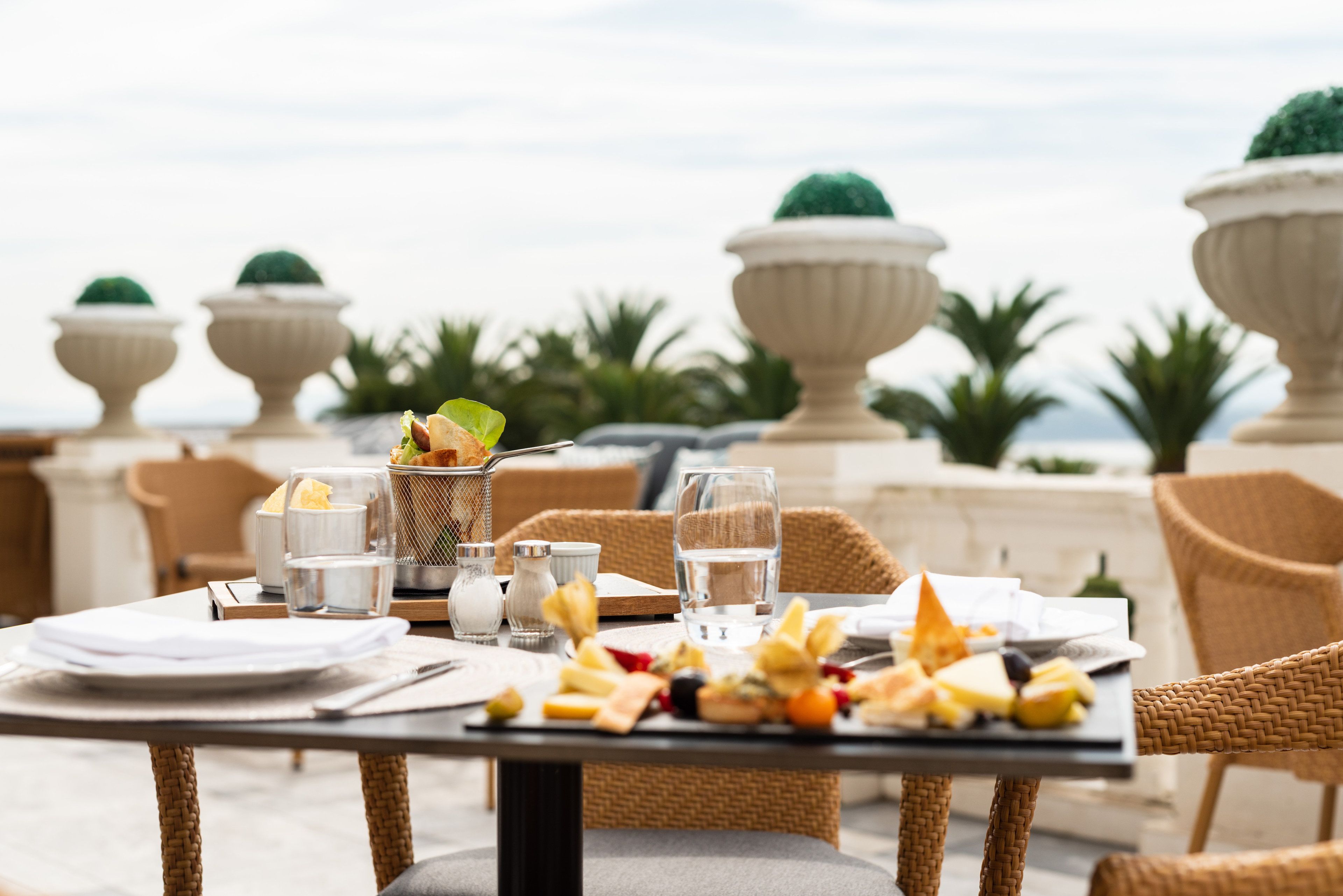 Mesa de comedor al aire libre con vista al mar, rodeada de sillas de mimbre y un jarro decorativo.