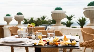 Mesa de comedor al aire libre con vista al mar, rodeada de sillas de mimbre y un jarro decorativo.