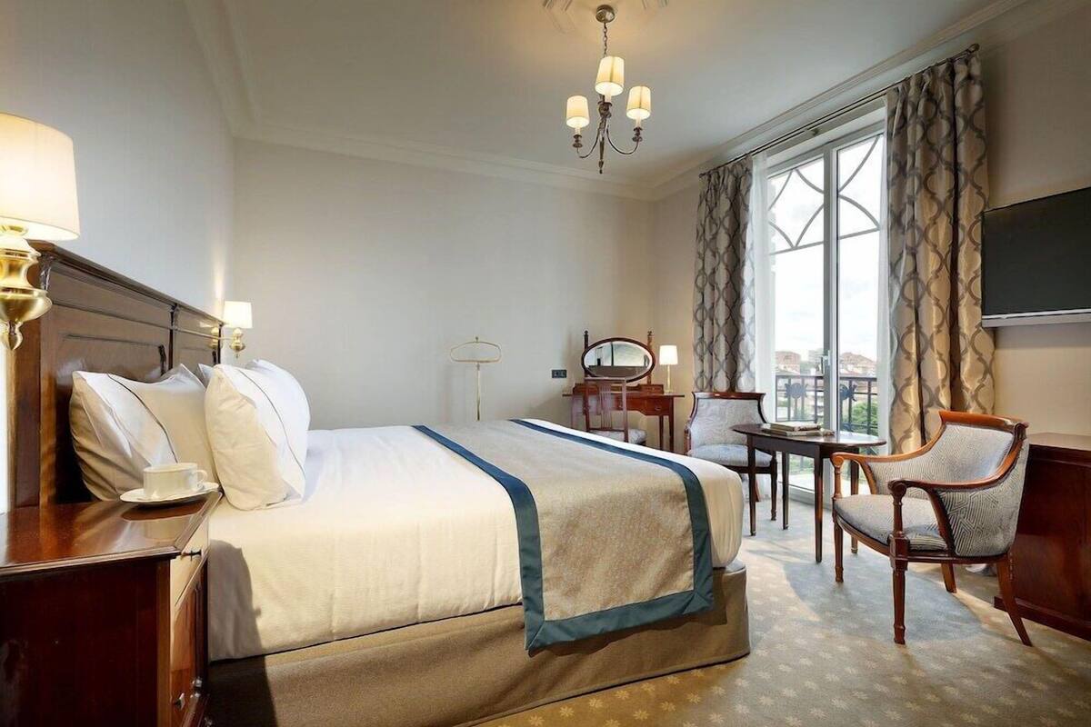 A hotel room with a large bed, a desk, a chair, a television, and a view of the city through the window.