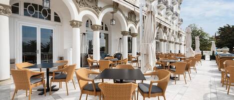 An outdoor café with wicker chairs and tables, a white building with ornate balconies, and a clear sky.