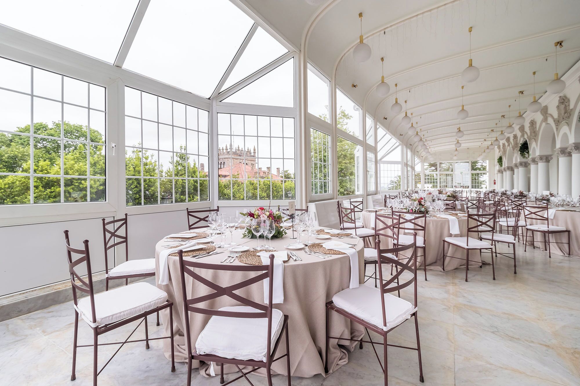 A spacious, elegantly decorated banquet hall with large windows offering a view of greenery and a distant building. Tables are set with white linens and centerpieces.