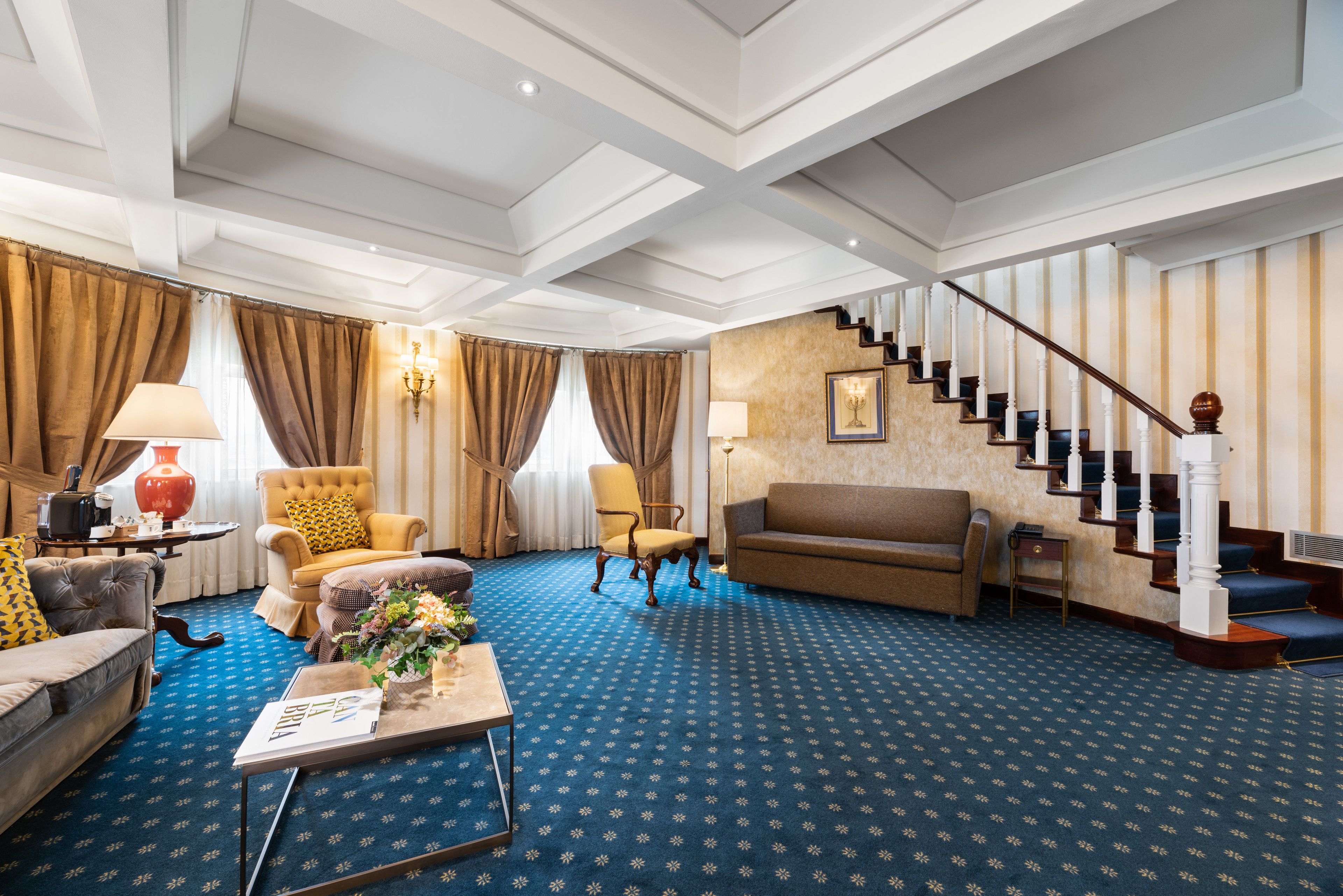 A spacious living room with a blue patterned carpet, a staircase with white balusters, and a chandelier.