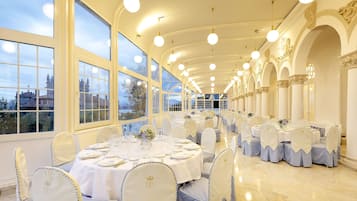 A banquet hall with round tables set for dining, large windows, and classical architecture.