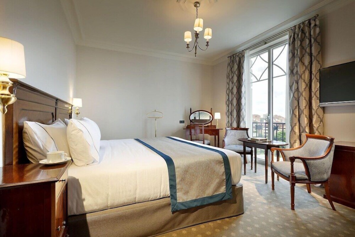 A hotel room with a large bed, a desk, a chair, a television, and a view of the city through the window.