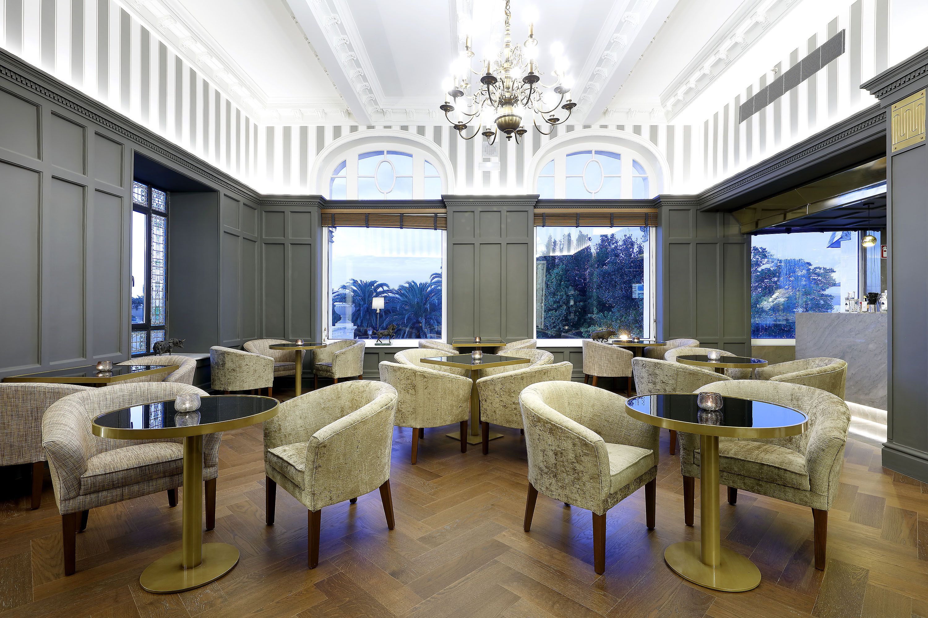 A room with multiple round tables and cushioned chairs, large windows with a view of trees, and a chandelier.