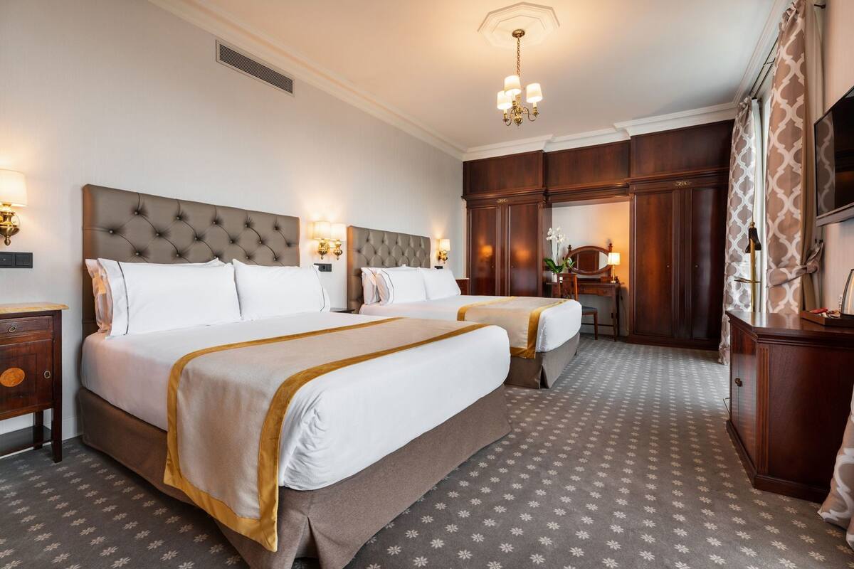 A hotel room with two beds, a desk, a wardrobe, and a chandelier.
