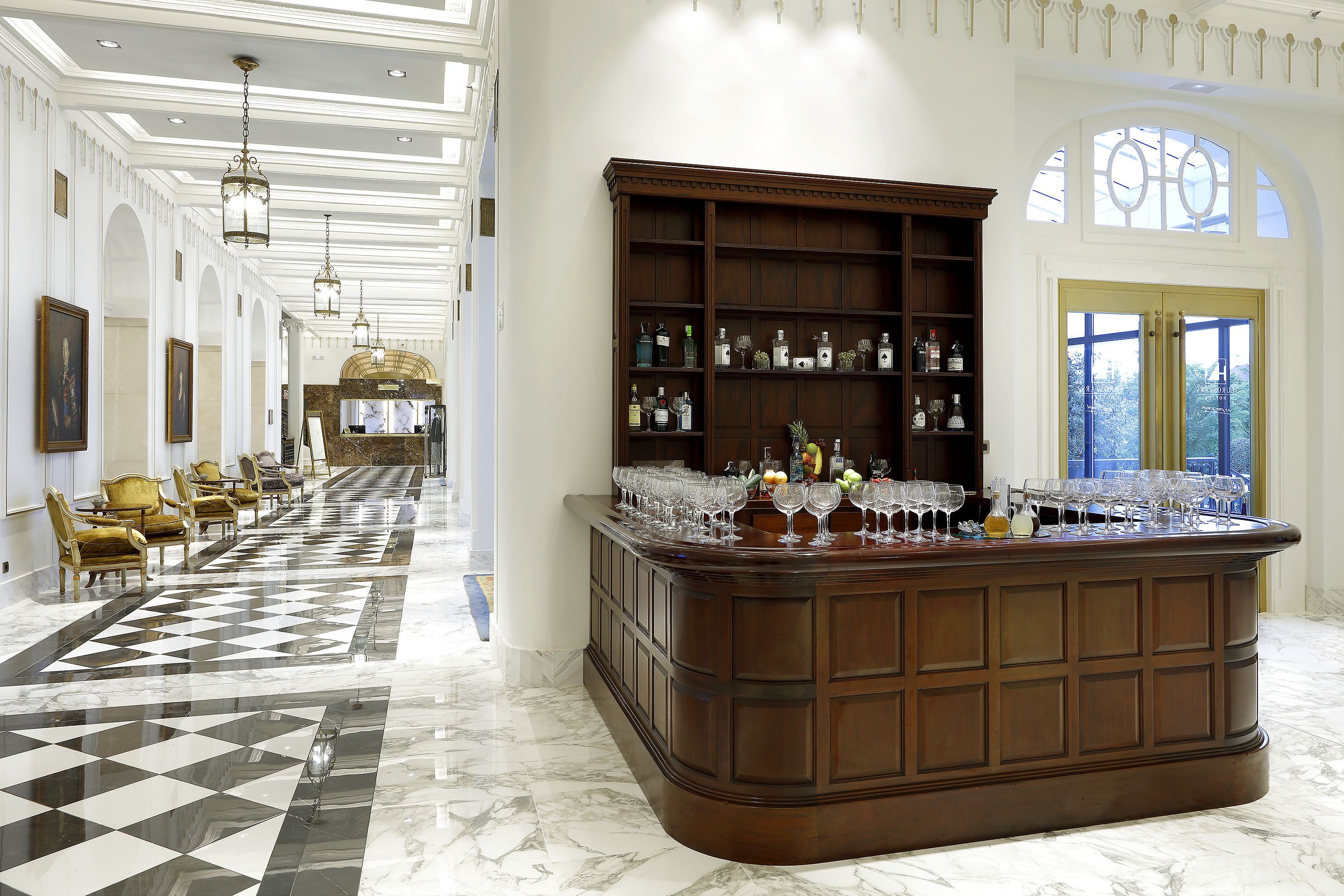 Um hall espaçoso com piso de mármore quadriculado, uma área de bar central com prateleiras de garrafas e copos, e um espaço de estar com poltronas estofadas.